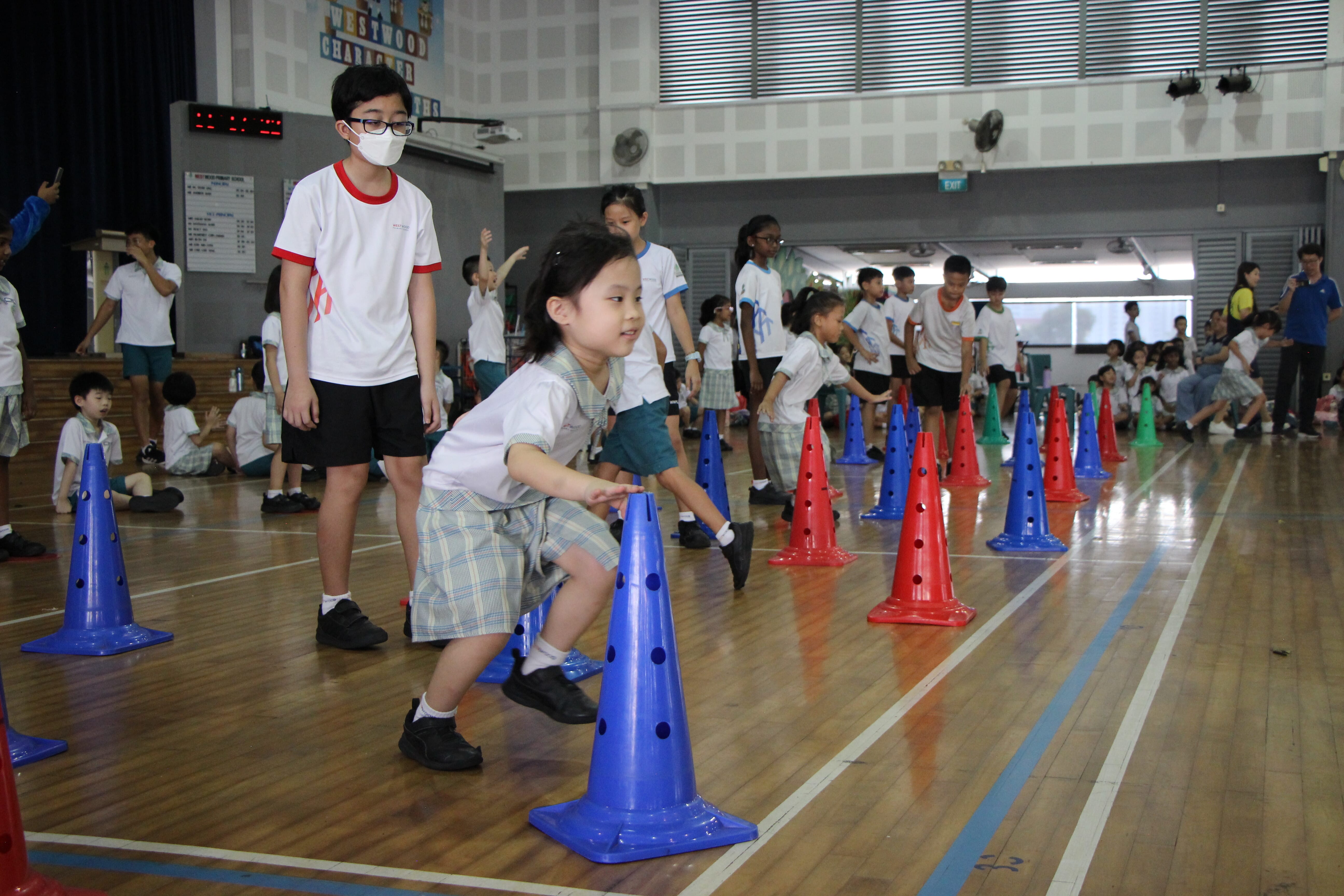 Children in school uniforms navigate blue and red plastic cones on a gymnasium floor during an indoor sports activity.